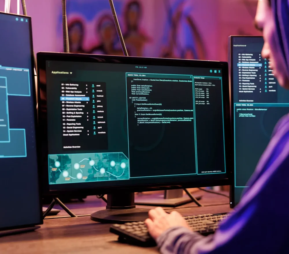 Hacker coding malicious software for password cracking in abandoned warehouse. Young man in hood programming illegal ransomware on computer with multiple monitors in hideout